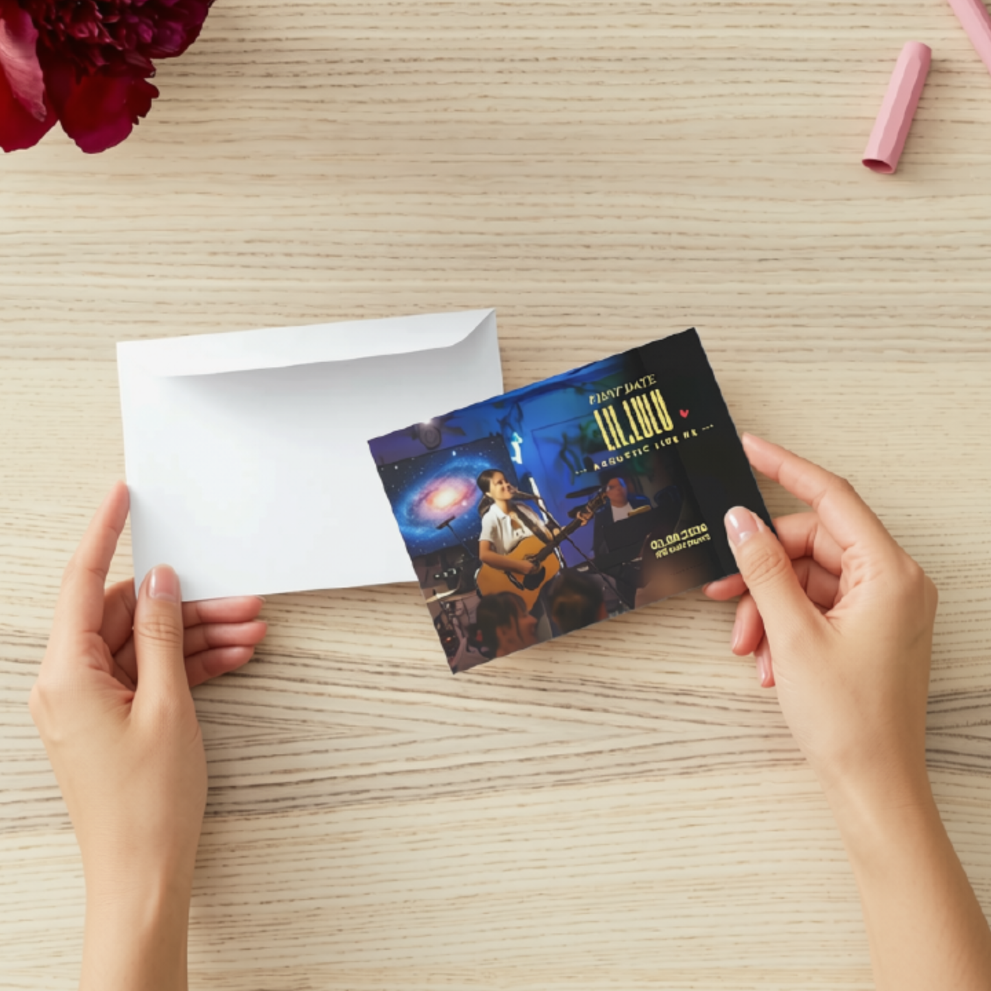 A person's hands hold a printed FIRST DATE with Lil.Lulu concert Postcard in front of a white envelope on a light wood tabletop. The card features a dark blue and black stage design with Lil.Lulu performing and the text FIRST DATE Lil.Lulu 02.08.2026